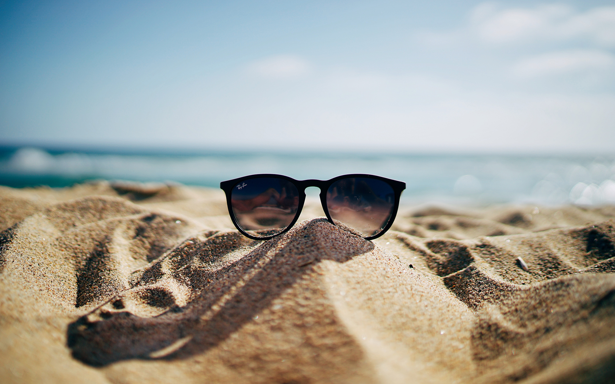 pair of sunglasses balanced on mound of sand with surf in background